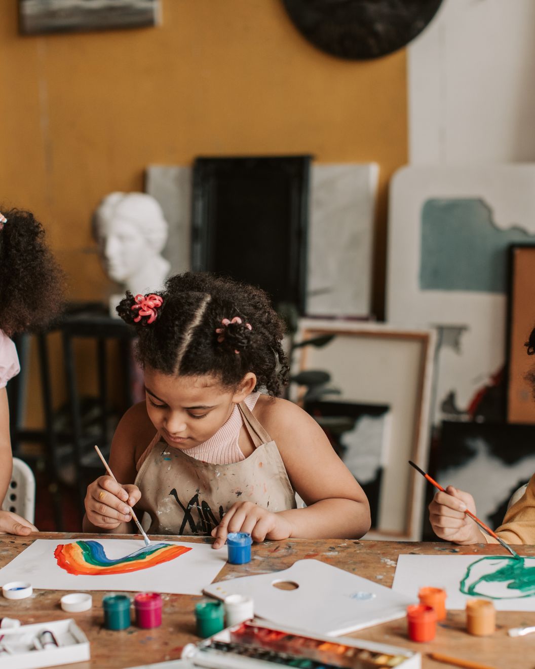 A young girl in an apron paints a rainbow at an art table with other children