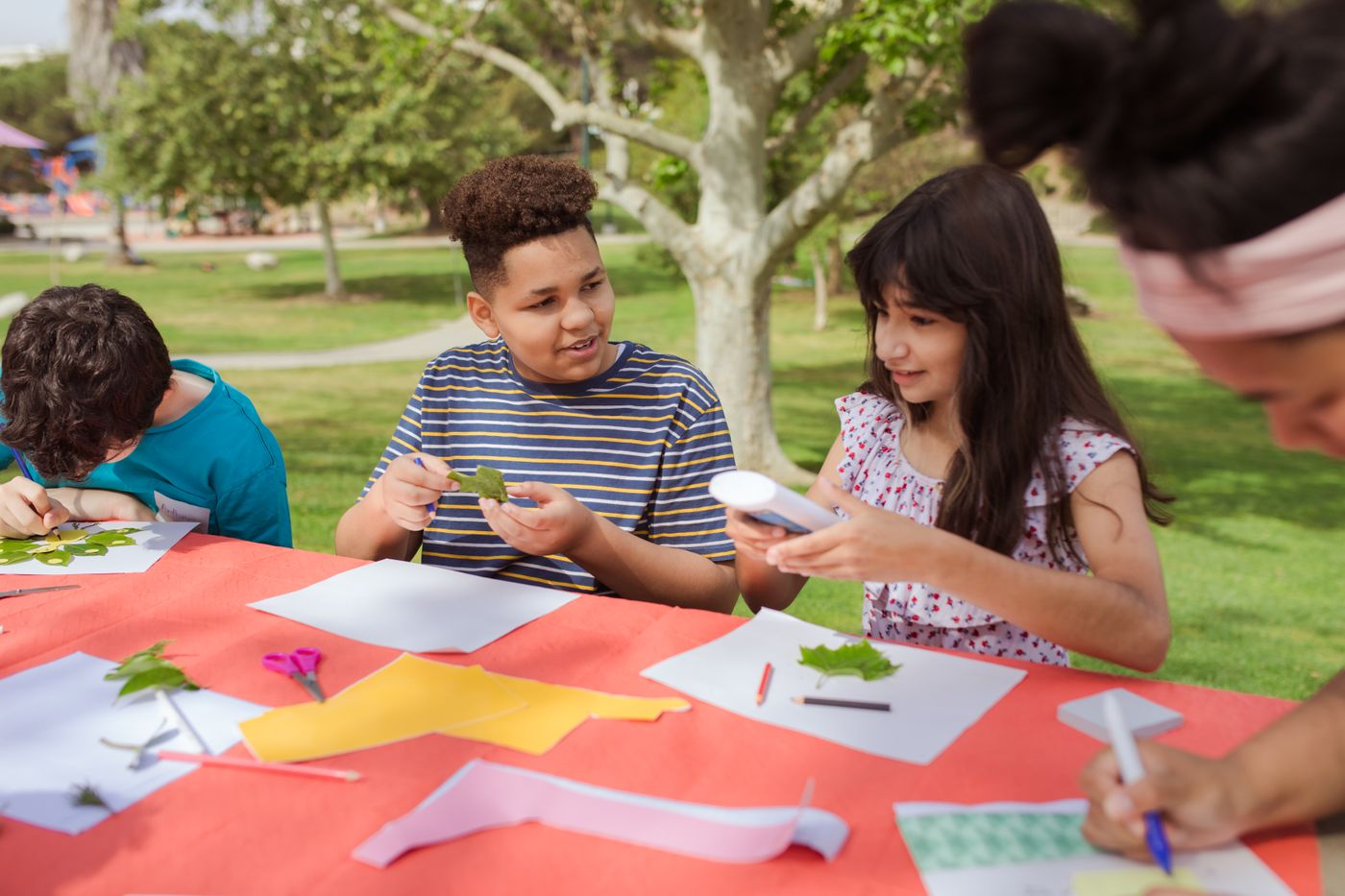Children make leaf collages together at a colorful table outdoors in a park