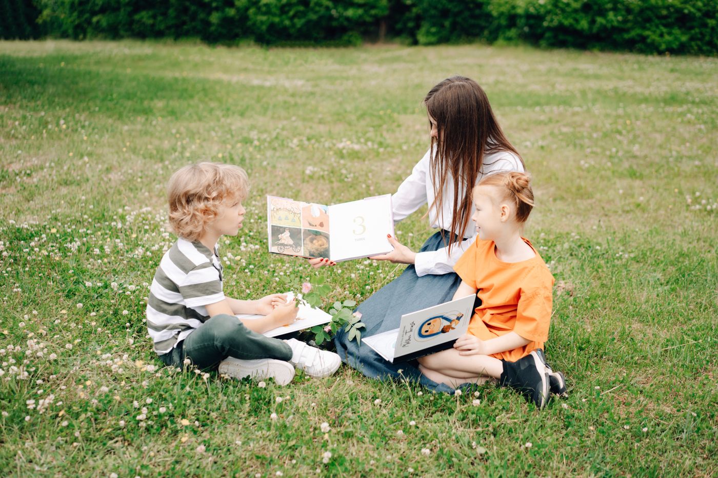 A teacher reads a picture book with two young children on the grass